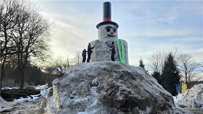 Großer Schneemann mit Zylinder und Karottennase auf einem Hügel aus Schnee, umgeben von mehreren Personen und Bäumen im Hintergrund.