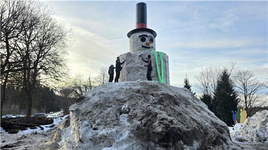 Großer Schneemann mit Zylinder und Karottennase auf einem Hügel aus Schnee, umgeben von mehreren Personen und Bäumen im Hintergrund.