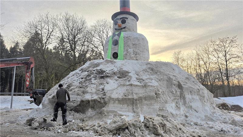 Großer Schneemann mit Zylinder und Karottennase auf einem Hügel aus Schnee, umgeben von mehreren Personen und Bäumen im Hintergrund.