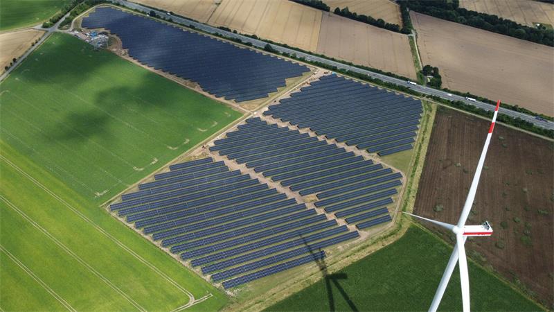 Ein Solarpark aus der Vogelperspektive, am oberen Bildrand verläuft eine Autobahn, rechts der Flügel eines Windrads.
