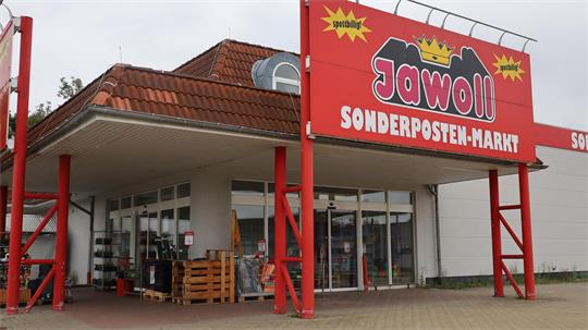 Eingangsbereich eines Gebäudes mit rotem Schild 'Jawoll SONDERPOSTEN-MARKT' auf weißer Fassade und rotem Dach