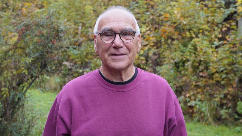 Älterer Mann mit Brille und lila Pullover steht draußen vor herbstlichem Gebüsch und lächelt.