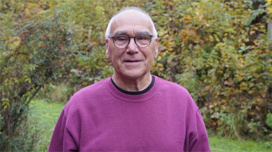 Älterer Mann mit Brille und lila Pullover steht draußen vor herbstlichem Gebüsch und lächelt.