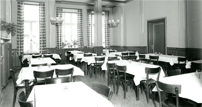 Speisesaal mit mehreren rechteckigen Tischen, die mit weißen Tischdecken bedeckt sind, und Holzstühlen in einem Raum mit hohen Fenstern und gemusterten Vorhängen