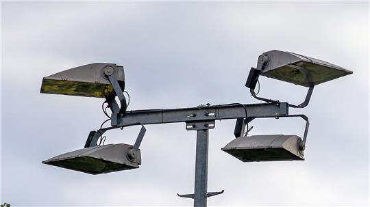Vier große, rechteckige Flutlichtstrahler an einem Metallmast vor bewölktem Himmel.