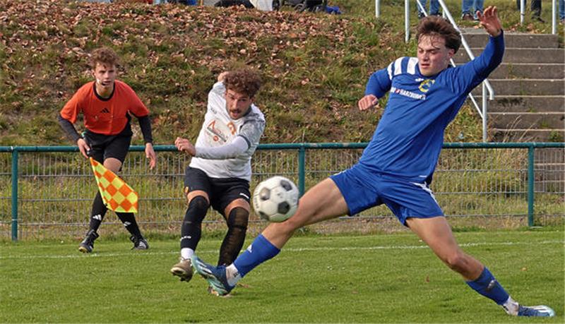 Zwei Fußballspieler in Aktion, einer in blau gleitet zum Ball, der andere in weiß steht bereit, im Hintergrund Schiedsrichter mit Fahne