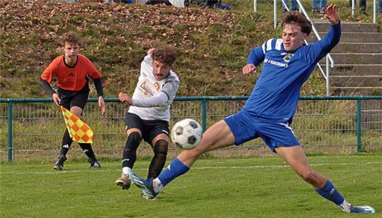 Zwei Fußballspieler in Aktion, einer in blau gleitet zum Ball, der andere in weiß steht bereit, im Hintergrund Schiedsrichter mit Fahne