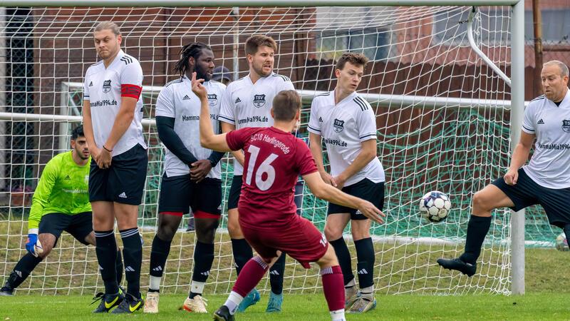 Fußballspieler in weißem Trikot bilden eine Mauer vor dem Tor, ein Spieler in rotem Trikot steht davor und bereitet einen Freistoß vor