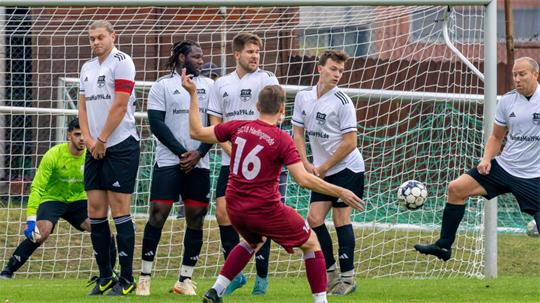 Fußballspieler in weißem Trikot bilden eine Mauer vor dem Tor, ein Spieler in rotem Trikot steht davor und bereitet einen Freistoß vor