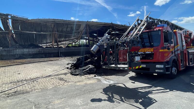Das Foto zeigt links einen seitlichen Blick auf die zerstörte Lagerhalle. Im Vordergrund steht rechts ein Feuerwehrfahrzeug, davor ist ein verkohlter Rest von Kunststoffpaletten zu sehen.
