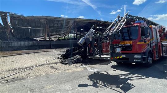 Das Foto zeigt links einen seitlichen Blick auf die zerstörte Lagerhalle. Im Vordergrund steht rechts ein Feuerwehrfahrzeug, davor ist ein verkohlter Rest von Kunststoffpaletten zu sehen.