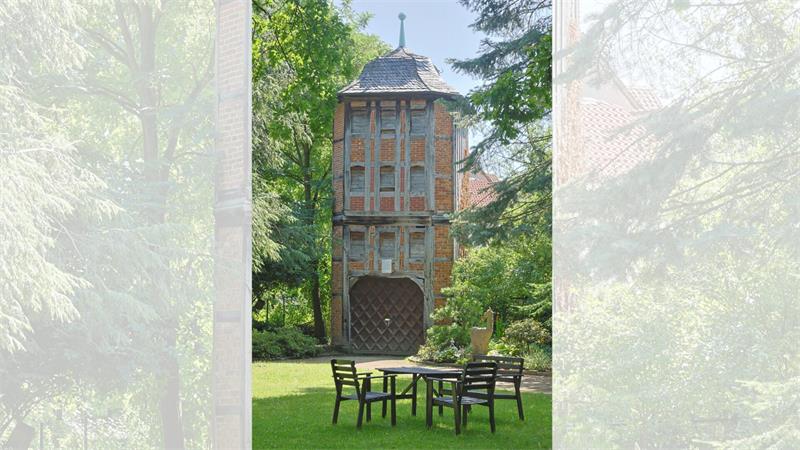 Fachwerkhaus mit spitzem Dach hinter einem Garten mit Tisch und vier Stühlen, umgeben von Bäumen
