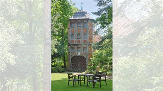 Fachwerkhaus mit spitzem Dach hinter einem Garten mit Tisch und vier Stühlen, umgeben von Bäumen