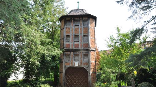 Ein hoher, schmaler Backsteinturm mit Holzverkleidung und einem spitzen Dach, umgeben von Bäumen und Grünflächen.