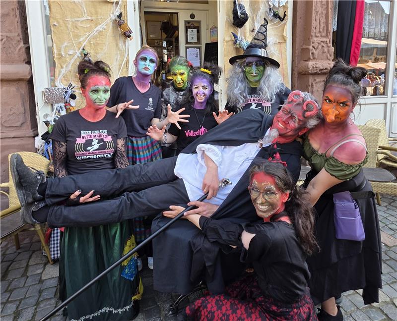 Gruppe von Menschen in historischen und fantasievollen Kostümen vor einem Café mit rotem Vorhang und Schild 'Cafe am Markt', eine Person mit rotem Teufelsgesicht wird von anderen getragen.