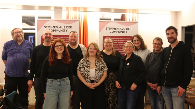 Gruppe von Menschen steht vor zwei Bannern mit der Aufschrift 'STIMMEN AUS DER COMMUNITY' in einem Raum mit Vorhängen und Spiegeln.