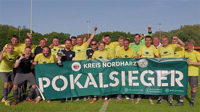 Gruppe von Fußballspielern in gelben Trikots steht auf dem Spielfeld und hält ein Banner mit der Aufschrift 'KREIS NORDHARZ POKALSIEGER'