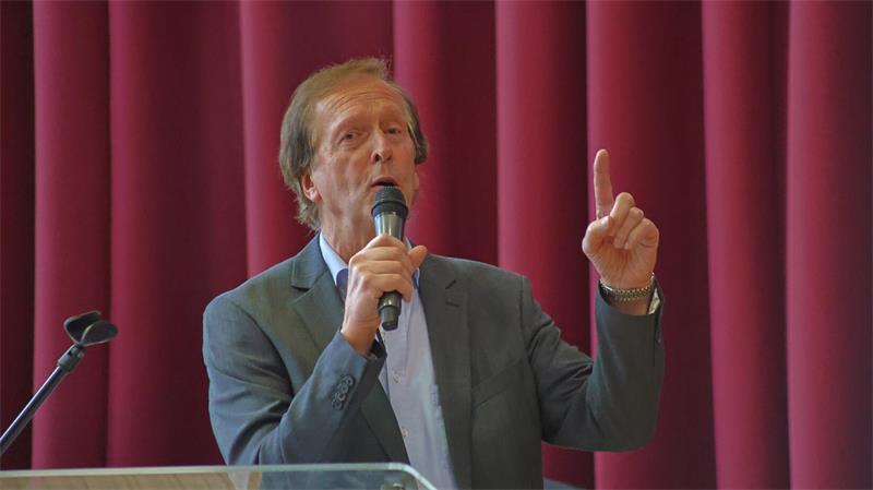Ein Mann in grauem Anzug spricht in ein Mikrofon und hebt den Zeigefinger seiner linken Hand vor einem roten Vorhang
