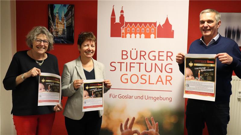 Der Vorstand der Bürgerstiftung Jutta Schober, Ulrike Olbrich und Meinhardt Fulst halten jeweils ein Poster in den Händen, das für die Spendenaktion „Leuchtende Kinderaugen“ wirbt. Auf dem Poster ist ein Bild eines kleinen Kindes zu sehen sowie Informationen zur Aktion.