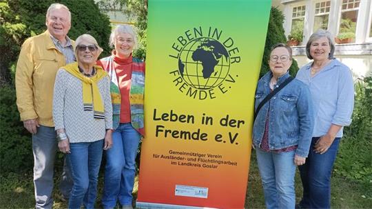 Fünf ältere Personen stehen vor einem Banner mit der Aufschrift 'Leben in der Fremde e.V.' und lächeln in die Kamera