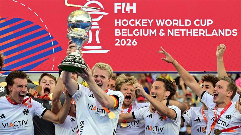 Männer-Hockeyteam jubelt, ein Spieler hält Pokal hoch, im Hintergrund Text 'FIH HOCKEY WORLD CUP BELGIUM & NETHERLANDS 2026'