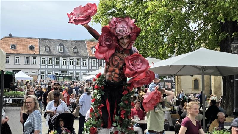 Person mit Blumenkostüm aus großen Rosen und grünen Blättern steht vor einer Menschenmenge auf einem Platz mit historischen Gebäuden und Sonnenschirmen.