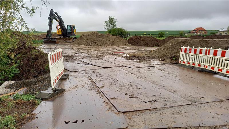 Baustelle mit Bagger, aufgeschütteten Erdhügeln und zwei Absperrungen auf matschigem Boden unter bewölktem Himmel.