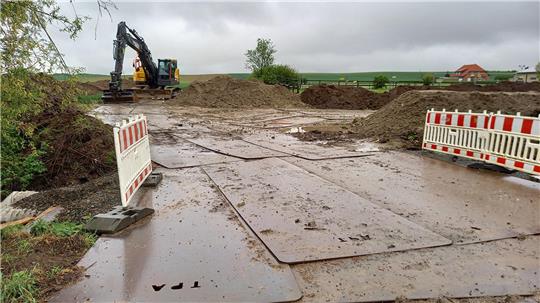Baustelle mit Bagger, aufgeschütteten Erdhügeln und zwei Absperrungen auf matschigem Boden unter bewölktem Himmel.