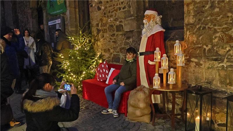 Kind sitzt auf roter Bank neben Weihnachtsmannfigur und beleuchtetem Weihnachtsbaum, Person fotografiert die Szene auf Kopfsteinpflaster vor Steinmauer