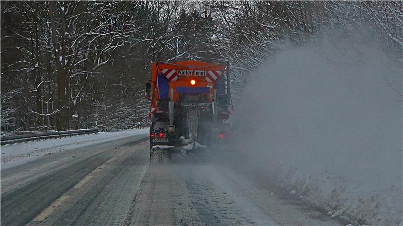 Streufahrzeug fährt auf schneebedeckter Straße durch bewaldete Winterlandschaft und verteilt Streugut