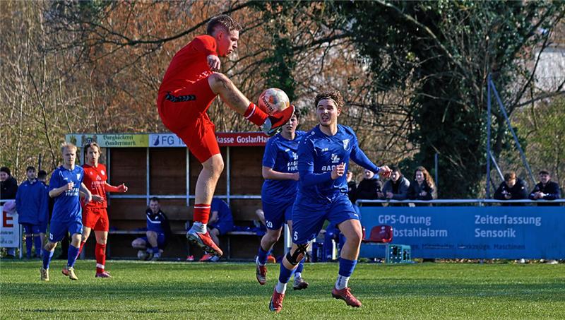 Fußballspieler in rotem Trikot springt und spielt den Ball mit dem Fuß gegen mehrere Spieler in blauen Trikots auf einem Spielfeld mit Zuschauern im Hintergrund