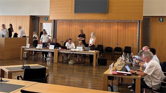 Mehrere Personen sitzen und stehen in einem großen Raum mit Holzwänden und Tischen, einige arbeiten an Laptops