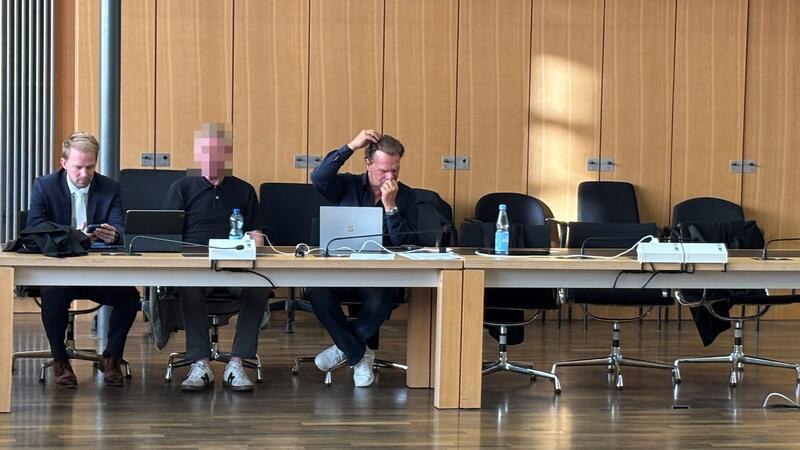 Drei Männer sitzen an einem langen Tisch, zwei von ihnen nutzen Laptops, einer schaut auf sein Smartphone, der mittlere Mann ist verpixelt.