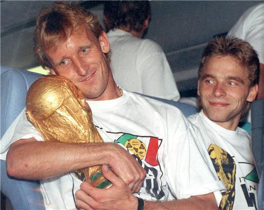 Der deutsche Abwehrspieler Andreas Brehme (l) hält 1990 in Rom den eroberten WM-Pokal im Arm und sein Teamgefährte Thomas Häßler schaut lächelnd zu (Archivbild vom 09.07.1990). Foto: picture-alliance / dpa | Frank_Kleefeldt
