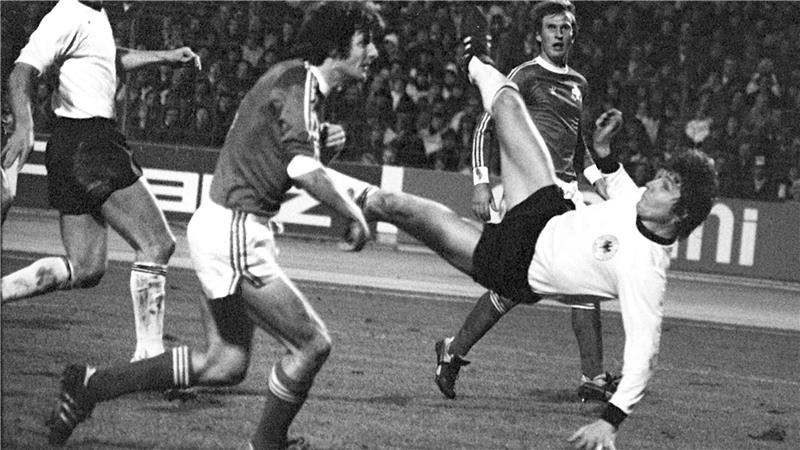 Fußballspieler in Aktion bei einem Spiel vor Zuschauern, einer führt einen Fallrückzieher aus