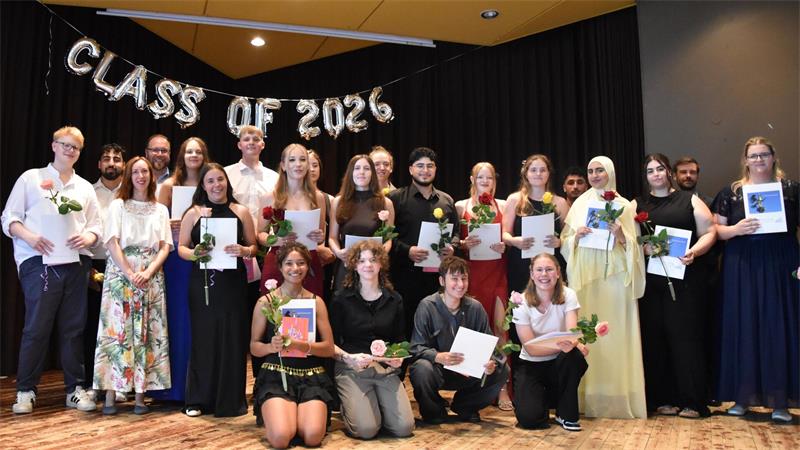 Gruppe von jungen Erwachsenen auf einer Bühne, einige halten Urkunden und Rosen, im Hintergrund silberne Luftballons mit der Aufschrift 'CLASS OF 2026'