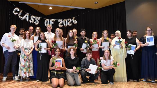 Gruppe von jungen Erwachsenen auf einer Bühne, einige halten Urkunden und Rosen, im Hintergrund silberne Luftballons mit der Aufschrift 'CLASS OF 2026'