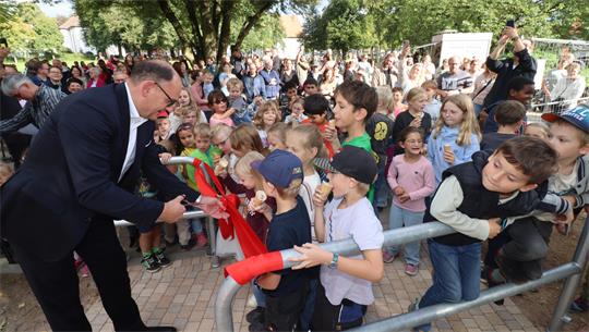 Ein Mann im Anzug durchschneidet mit einer Schere ein rotes Band, viele Kinder stehen an.