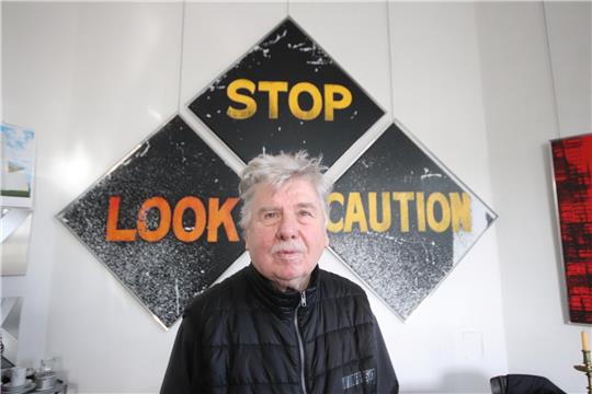 Das Foto zeigt Gerd Winner in seinem Atelier. Im Hintergrund sind drei quadratische Kunstwerke zu sehen, die  wie ein Pfeil angeordnet sind. Die Kunstwerke zeigen die Wörter "Stop", "Look" und "Caution" in gelber Schrift auf schwarzem Untergrund.