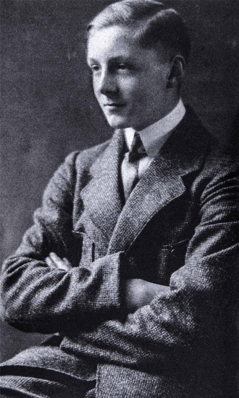 Ein junger Mann in Anzug mit verschränkten Armen sitzt vor dunklem Hintergrund.