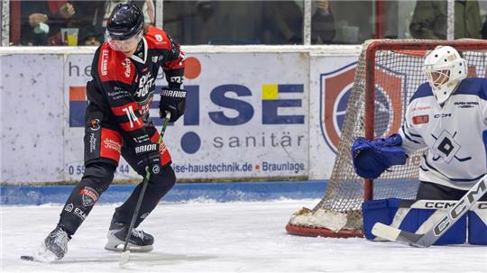 Eishockeyspieler in schwarzer und roter Ausrüstung mit Schläger vor dem Tor, Torwart in weißer Ausrüstung in Bereitschaftshaltung.