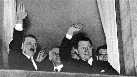 Vier Männer in Anzügen winken aus einem Fenster mit weißen Vorhängen.