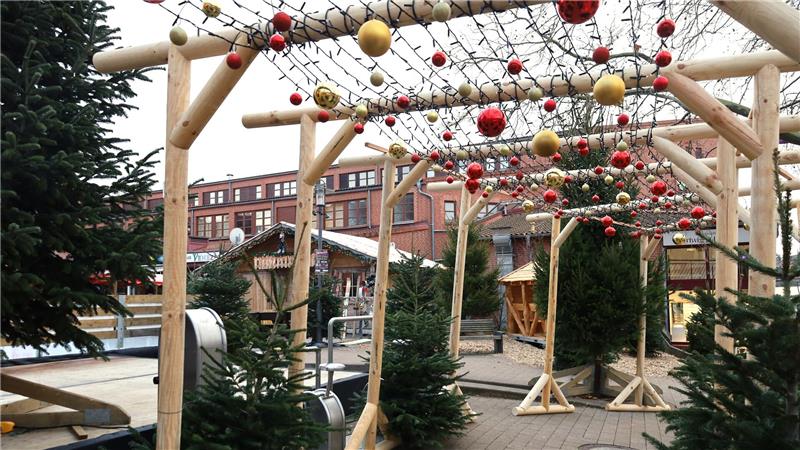 Kugeln und Lichterketten sind an einer Holzkonstruktion befestigt, durch die man durchgehen kann. Drumherum stehen Tannen.