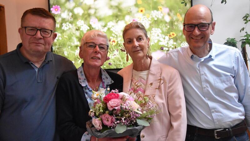 Vier Personen stehen eng beieinander, zwei Männer außen, zwei Frauen in der Mitte mit einem Blumenstrauß.
