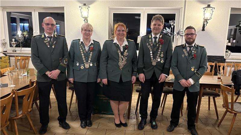 Fünf Personen in grünen Uniformjacken mit Orden stehen in einem Raum mit Holztischen und Stühlen vor einer weißen Wand mit zwei Wandlampen