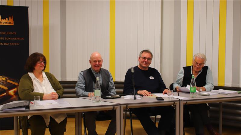 Vier Personen sitzen an einem Tisch mit Mikrofon und Wasserflaschen vor einem weißen Hintergrund mit gelben Streifen