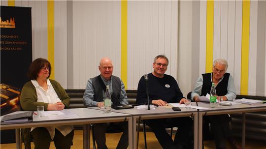 Vier Personen sitzen an einem Tisch mit Mikrofon und Wasserflaschen vor einem weißen Hintergrund mit gelben Streifen