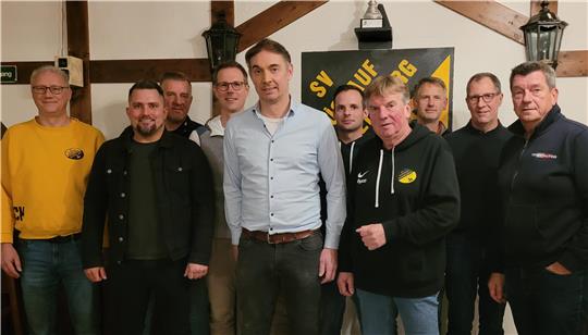 Gruppe von neun Personen steht in einem Raum mit Holzbalken und einem Schild mit gelbem und schwarzem Logo an der Wand.
