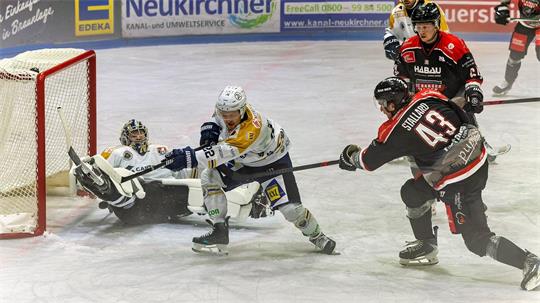 Ein Eishockeyspieler in schwarzer Spielkleidung schießt, zwei Spieler in weißer Kleidung versuchen, den Puck aufzuhalten.