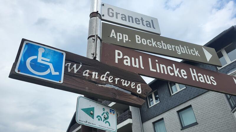 Wegweiser mit den Aufschriften 'Granetal', 'App. Bocksbergblick', 'Paul Lincke Haus' und einem Rollstuhlsymbol mit dem Wort 'Wanderweg' vor einem mehrstöckigen Gebäude und bewölktem Himmel